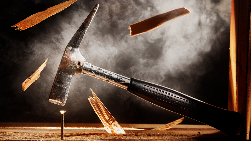 Holzsplitter fliegen druch die Luft, während ein Hammer auf einen Nagel einschlägt.