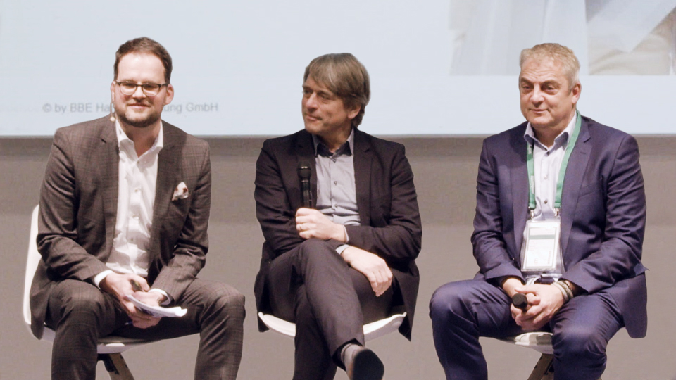 Sebastian Deppe, Martin Kremming und Daniel Schnödt diskutieren auf einer Bühne vor einer Projektion.