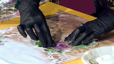 Black-gloved hands apply a rice paper rose on a textile bag.
