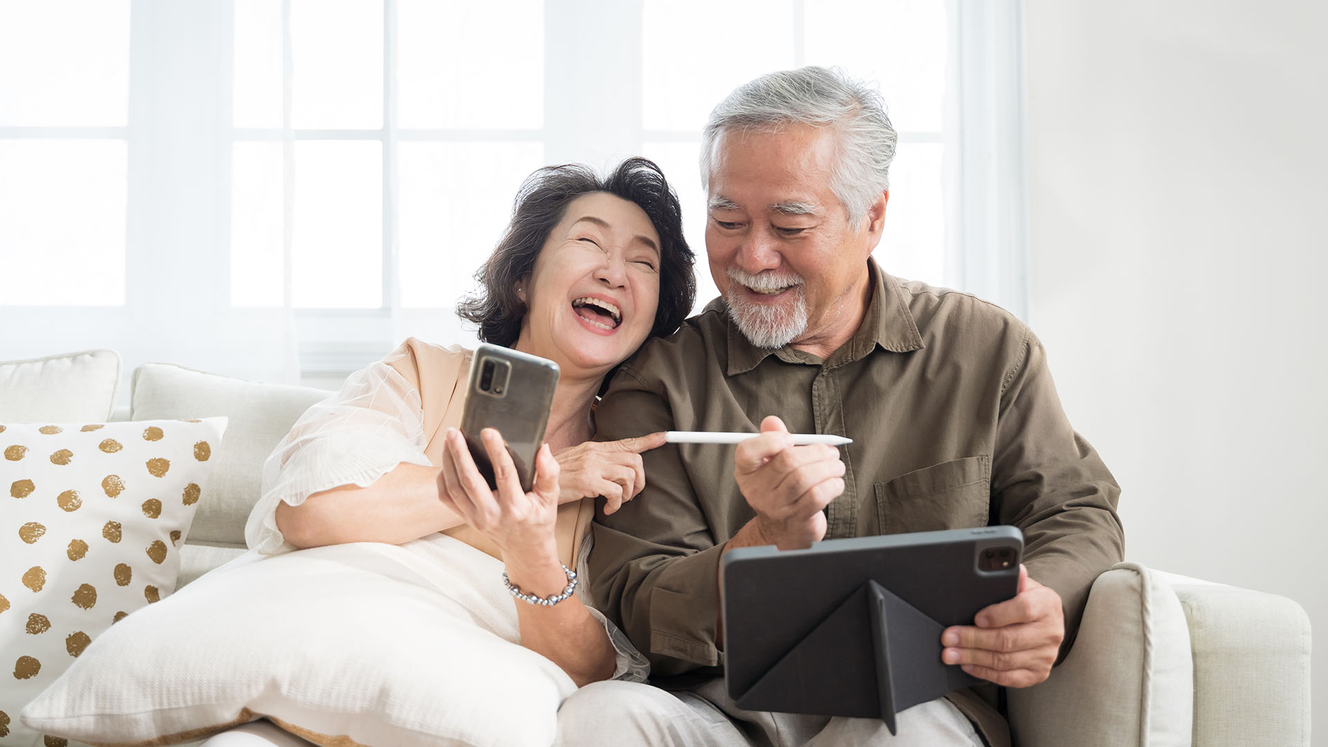 An elderly Asian couple on smartphones and tablets, laughing together